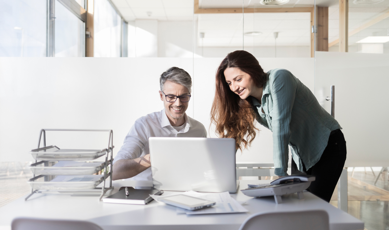 A woman stands next to a seated man and they both look at a laptop in an office setting