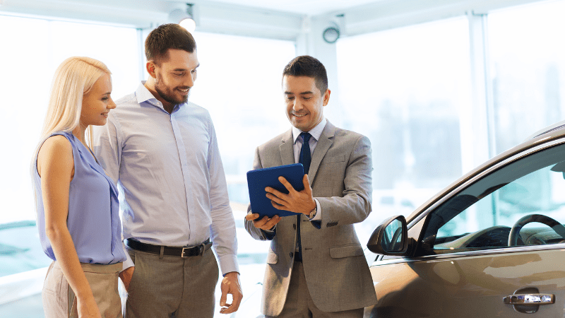 A man on the right of the photo stood next to a car shows a couple something on his tablet device whilst stood in a car showroom.
