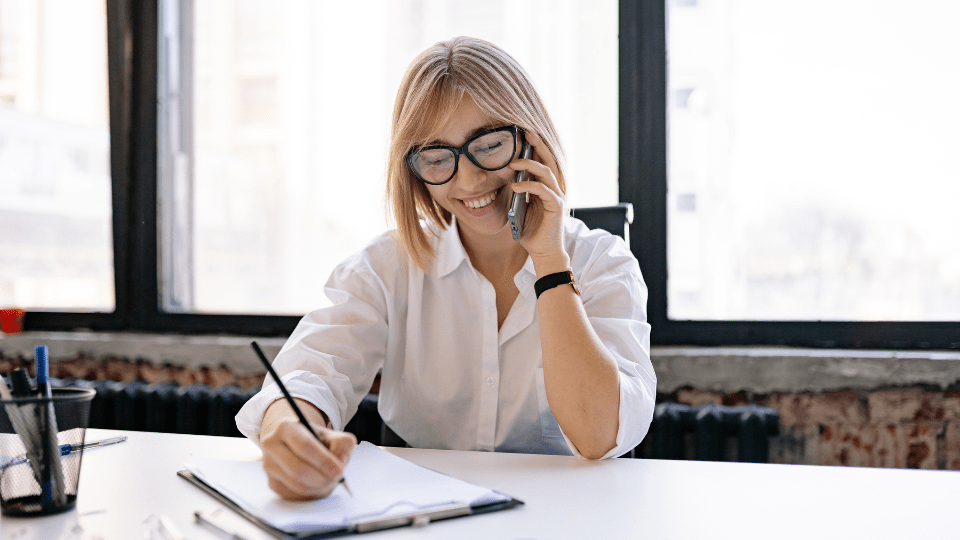 Woman in a white shirt with black glasses and blonde hair holding a phone to her ear whilst writing on a notepad situated on a table in front of her.
