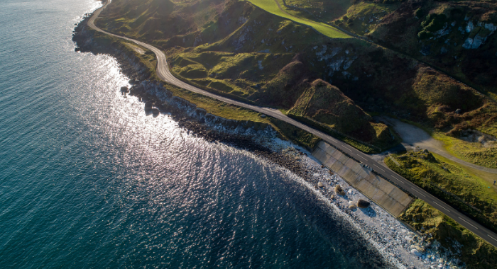 Causeway coastal An arial image of the Causeway Coastal. The sea takes up the left side of the image with the road on the coast and hills built up top the right of it.
