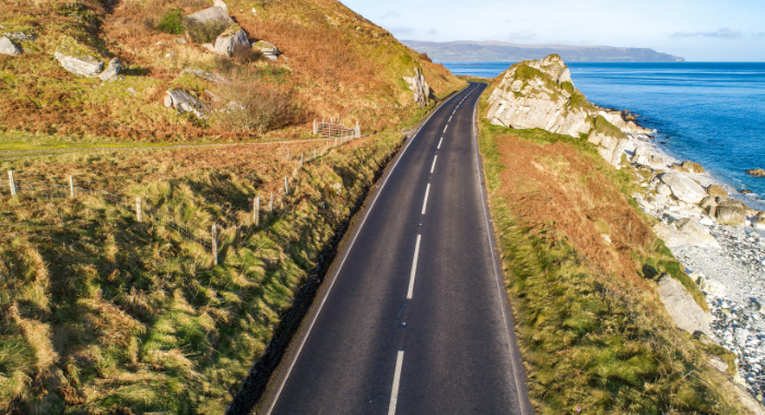 An arial view of the Causeway Coastal Highway. The road is central in the image with a grassy hill to the left and a cliff drop to the sea on the right.