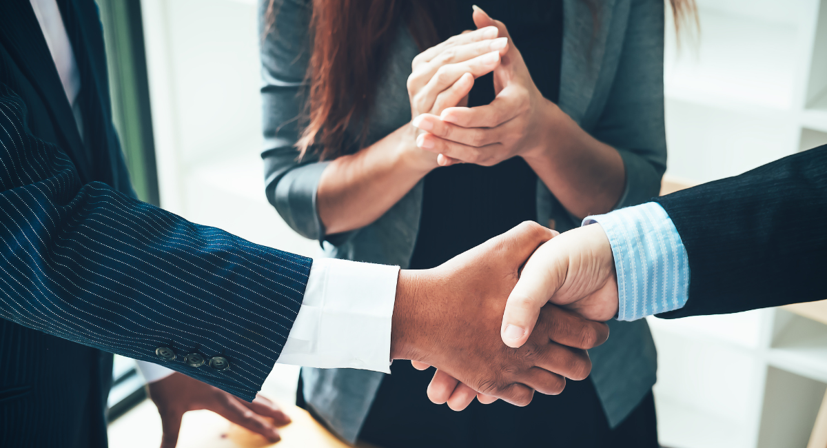 Commitment Two business people shake hands with a woman stood in the background with her hands together about to clap.