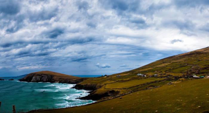 Dingle Brown coloured hills from the right slope down to meet the sea on the left, causing breaks and waves. A blue cloudy sky sits in the background.