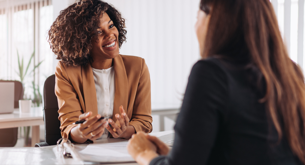 Two women sit across from one another with one having her back turned to the camera. They are engaged in a conversation and the woman facing the camera is smiling.