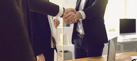 Two business professionals shaking hands whilst another person watches on.
