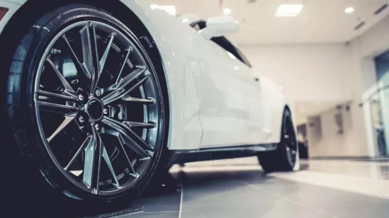 A white car in a showroom with black wheels and dark grey rims, with a lit ceiling in the background and the camera taken from eye level with the front right tyre looking back along the car.