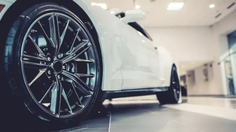 new-car-wheel-low A white car in a showroom with black wheels and dark grey rims, with a lit ceiling in the background and the camera taken from eye level with the front right tyre looking back along the car.