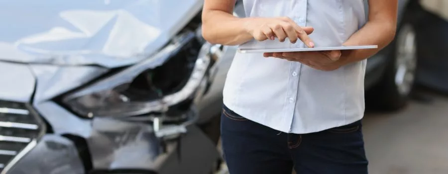 A person in a white shirt stands in front of a parked car whilst scrolling on a tablet device.