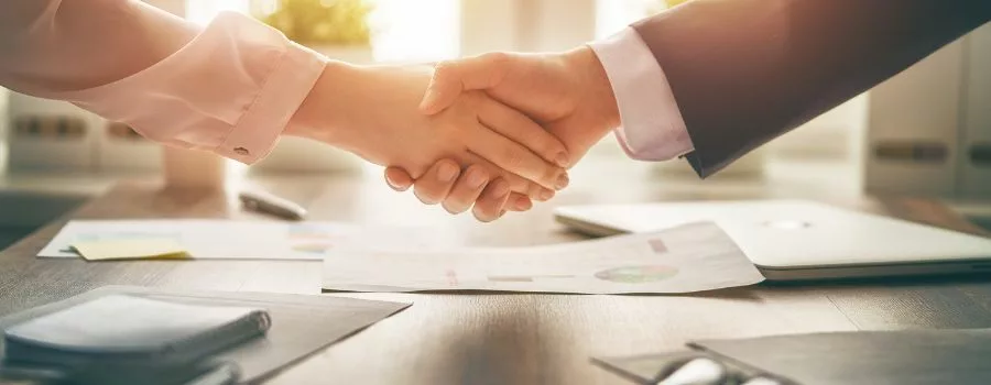 Two people shake hands over a table with documents on.