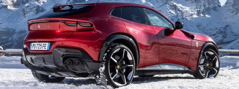 A red sports coupe is parked on snow with a backdrop of rugged snow-covered mountains under a clear blue sky.