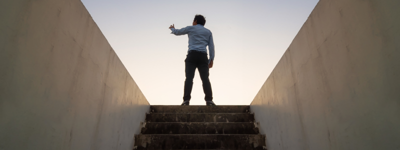 <br />
A man standing at the top of a staircase, raising his hand towards the sky, with a clear sky in the backdrop, depicting a concept of success or aspiration.