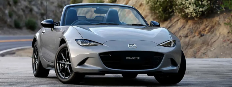 A grey Mazda MX5 roadster convertible on the side of a road with hills rising to the side of it.