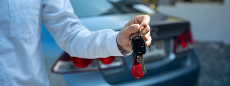 A person hands over car keys with a car behind them.