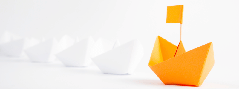 White origami pieces in a line, leading to an orange version with an orange flag on top.
