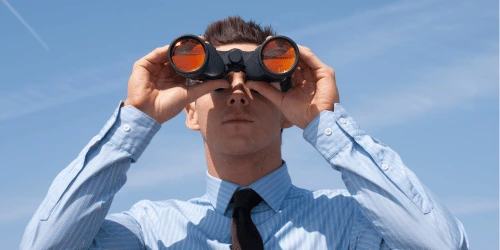 A person in a shirt and tie looking forward with binoculars.