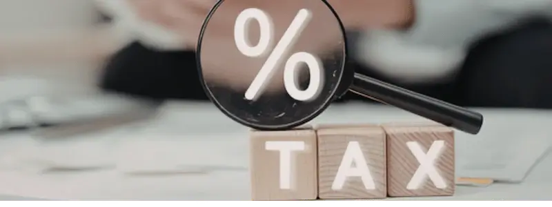 A magnifying glass focusing on a percentage sign with 'TAX' letter blocks in the foreground.